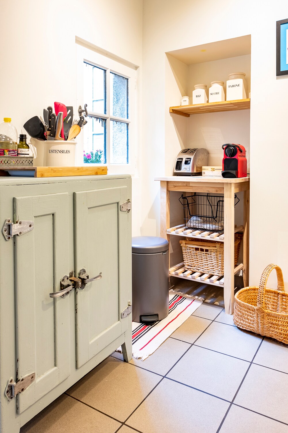 A functional kitchen nook is presented, featuring a mint-green cabinet and a wooden shelf holding a coffee machine and storage containers. A basket and trash bin are noticeably arranged below. Natural light streams through the nearby window, illuminating the tiled floor.