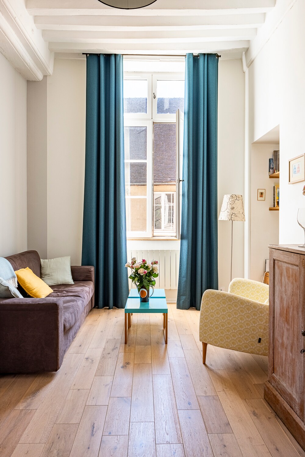 The living space features a brown sofa and a yellow-patterned chair, oriented towards a glass table. Light floods in through large windows dressed in blue curtains, highlighting the wood flooring and creating a peaceful ambiance.