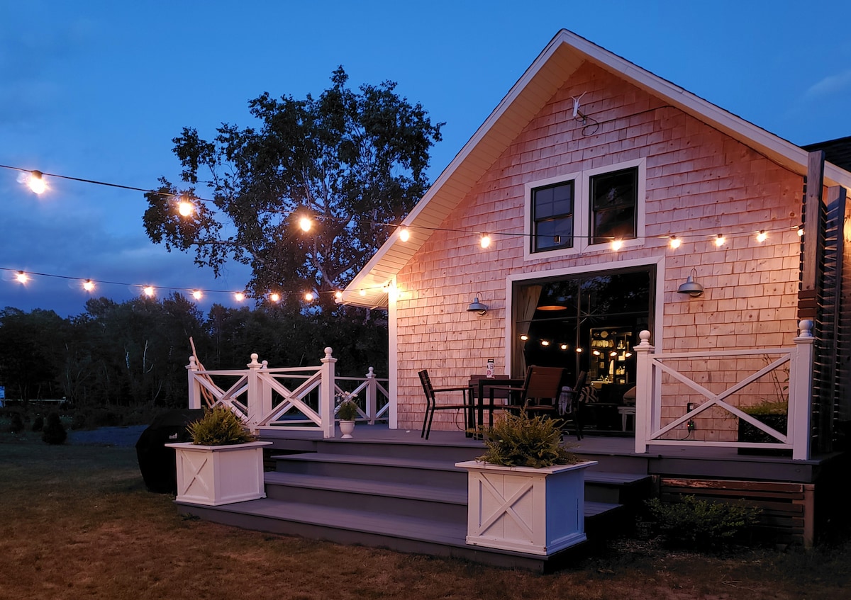 The cottage exterior features a charming wood shingle facade, with a welcoming deck adorned with string lights. The entrance is framed by large glass doors, leading into the inviting interior, while potted plants add a touch of greenery on the deck.