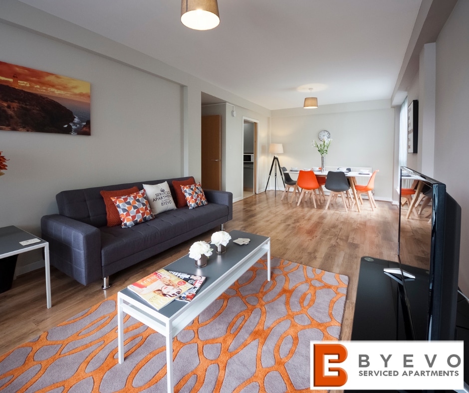 A modern living area features a dark sofa adorned with colorful cushions and a coffee table with decorative items. A vibrant area rug adds warmth to the space, which is complemented by a dining area with chairs in various colors. Natural light filters in through the large window.