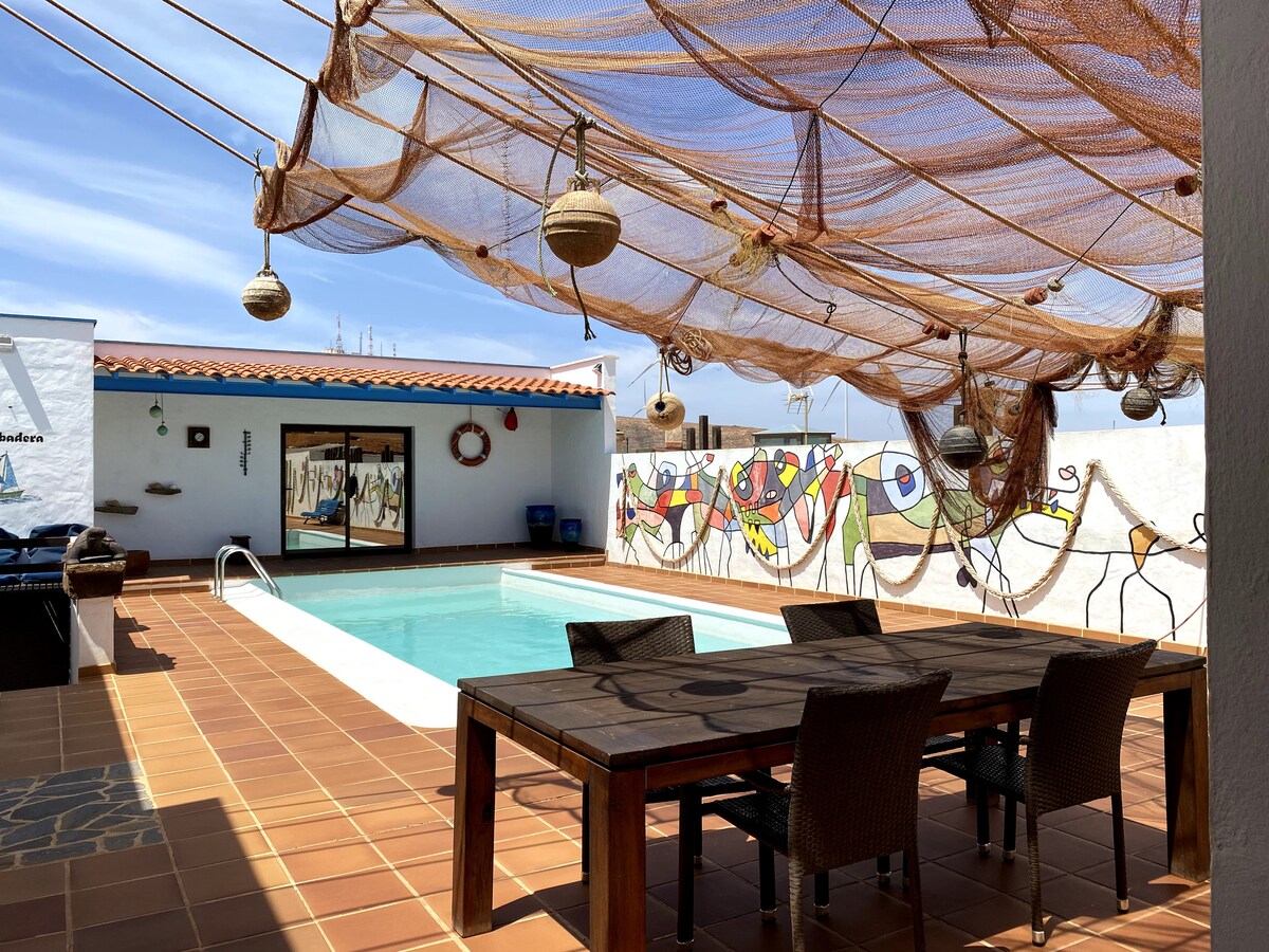 An outdoor area features a private swimming pool surrounded by tiled flooring. A large wooden table with woven chairs is placed under a decorative canopy, offering shade. Colorful murals adorn the white walls, enhancing the space's vibrant atmosphere.