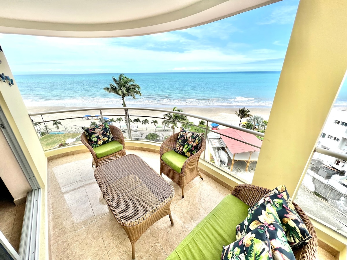 A covered balcony features comfortable wicker seating with vibrant floral cushions. A low table is positioned at the center, offering unobstructed views of the ocean and shoreline. Palm trees frame the scene, while the calm waters extend into the horizon under a clear sky.