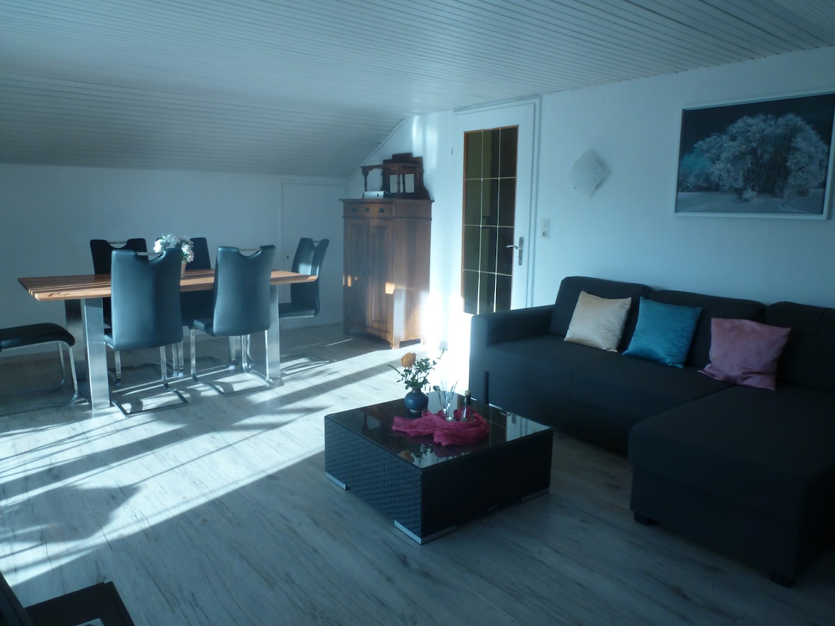 A well-lit living and dining area features a modern dining table with six black chairs and a dark sectional sofa. A coffee table with decorative items sits in front of the sofa. Natural light streams in through the windows, enhancing the spacious atmosphere.