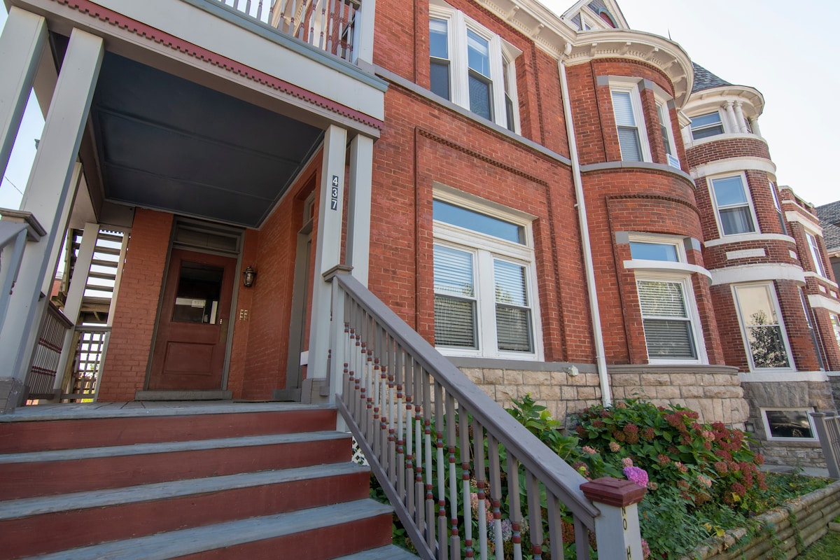 The exterior of the Victorian mansion showcases classic red brick with beautiful architectural details. A set of stairs leads to the main entrance, adorned with a wooden railing. Lush greenery and flowers enhance the inviting front garden.