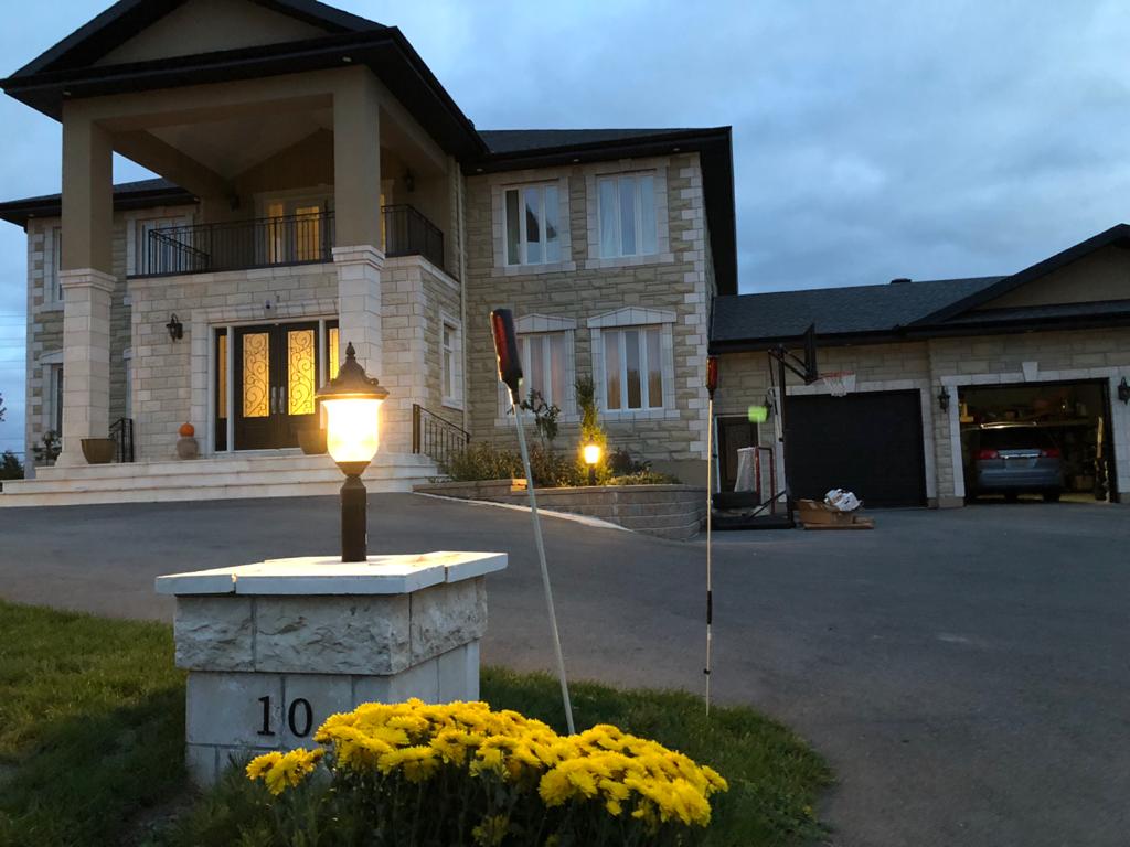 A spacious two-story home is pictured, featuring a prominent entryway with decorative pillars. Soft outdoor lighting highlights the stone façade. Yellow flowers are arranged at the base of a lamp post, enhancing the inviting entrance. A driveway leads to a garage on the right.