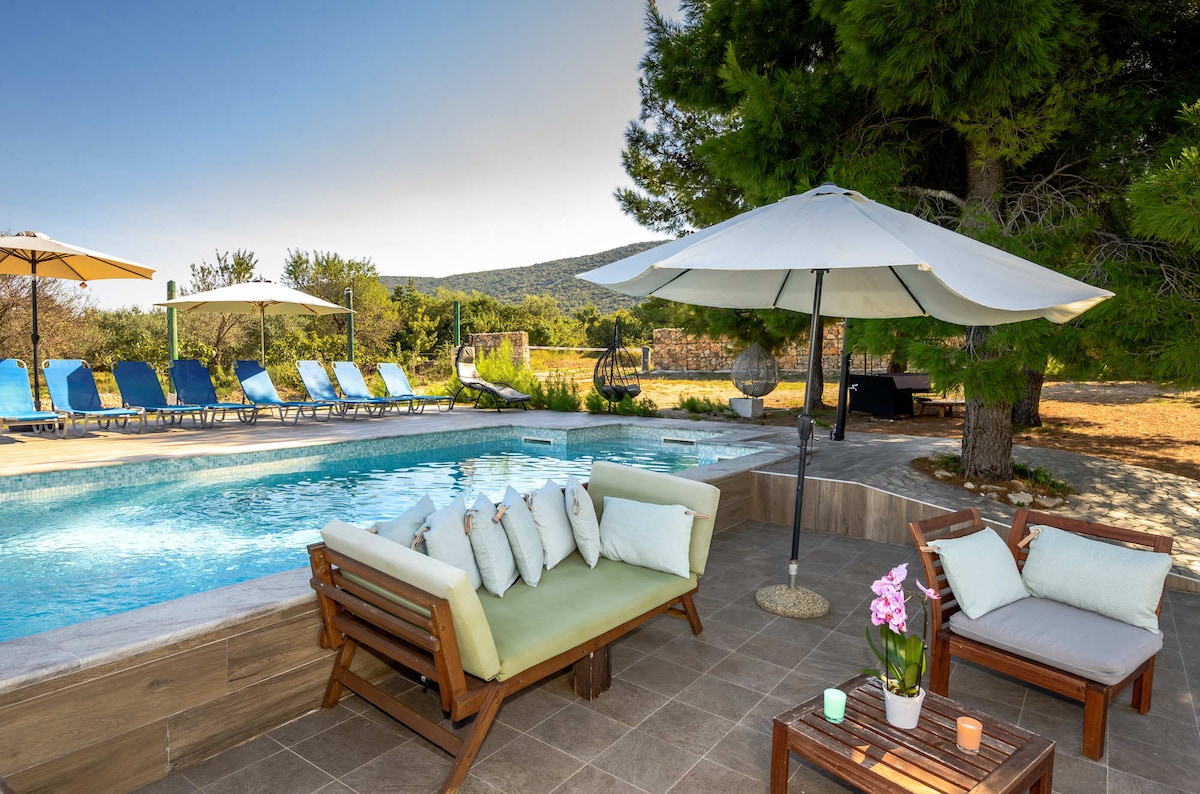 A serene outdoor area features a refreshing swimming pool surrounded by sun loungers and shaded umbrellas. Comfortable seating is arranged nearby, adorned with decorative cushions. Lush greenery frames the space, providing a sense of privacy and tranquility.