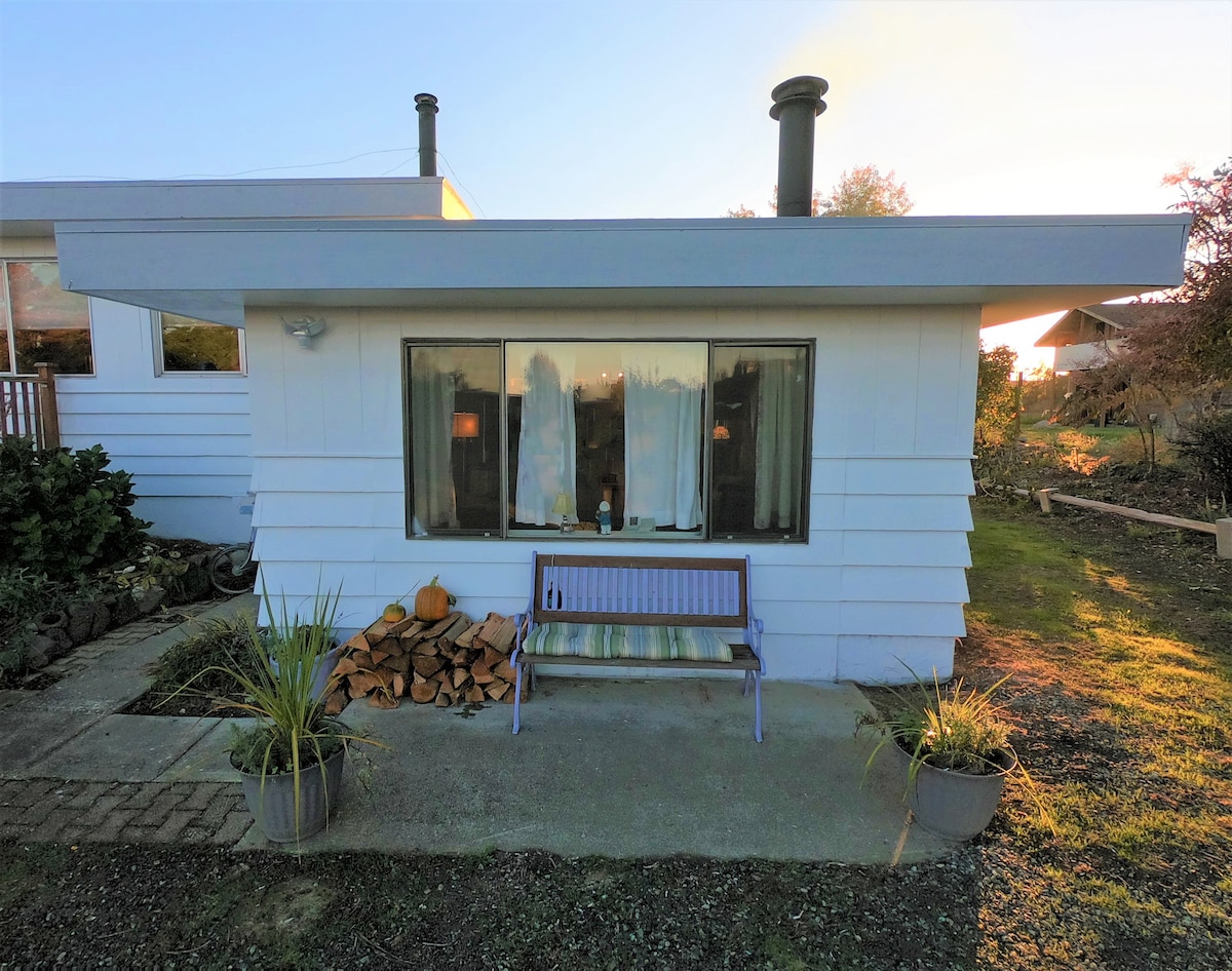 The exterior of the cottage is presented, featuring a covered porch with a purple bench. A stack of firewood is neatly arranged beside the cottage, and flower pots are placed on either side of the entrance. Large windows allow natural light to fill the space.