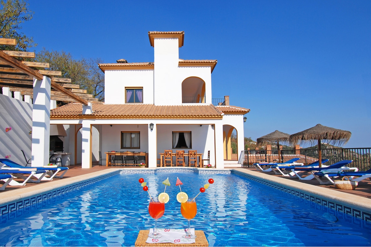 The villa's exterior features a well-maintained private heated swimming pool, flanked by loungers and shaded areas. A spacious terrace is visible, furnished with a dining set under a pergola. The bright architectural design of the villa stands against a clear blue sky.