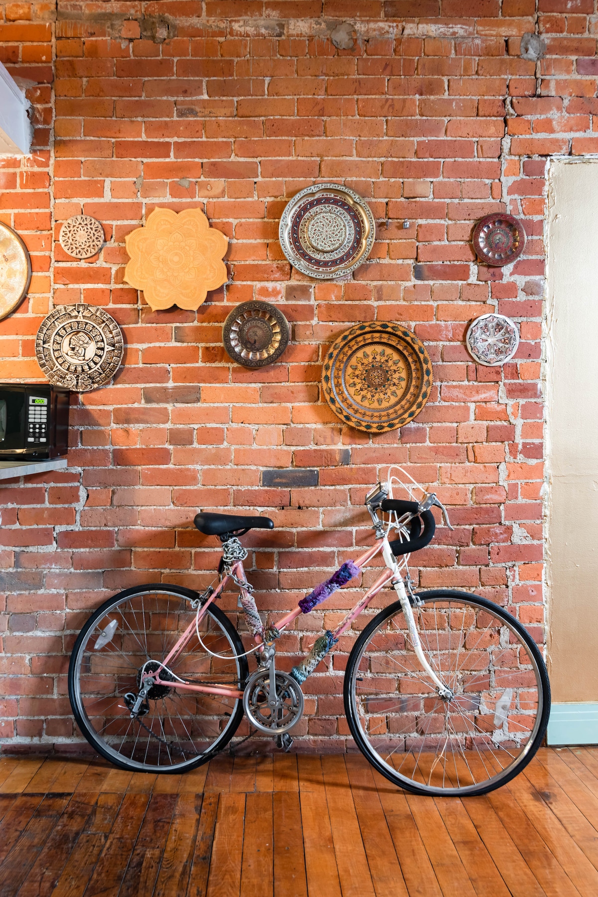 A vintage bicycle is positioned against a brick wall adorned with various decorative plates, adding character to the loft's interior. The warm wood floors contrast with the texture of the exposed brick, contributing to the industrial aesthetic.