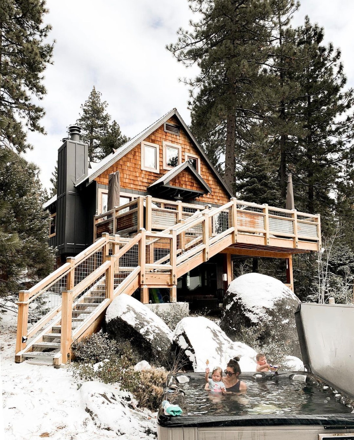 A charming cabin is set against a backdrop of pine trees and snow-covered boulders. A hot tub is positioned on a deck, with steam gently rising. Stairs lead up to the entry, which is framed by wooden railings, showcasing rustic architecture.