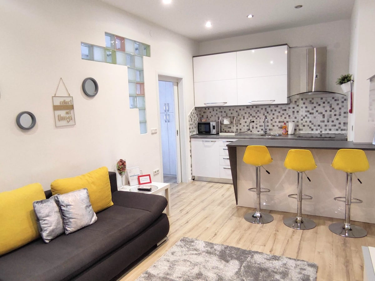 A modern kitchen features sleek white cabinetry and a patterned backsplash. Yellow bar stools are positioned at a small counter, complementing a cozy living area with a dark sofa adorned with decorative pillows. Light wood flooring enhances the spacious feel of the room.