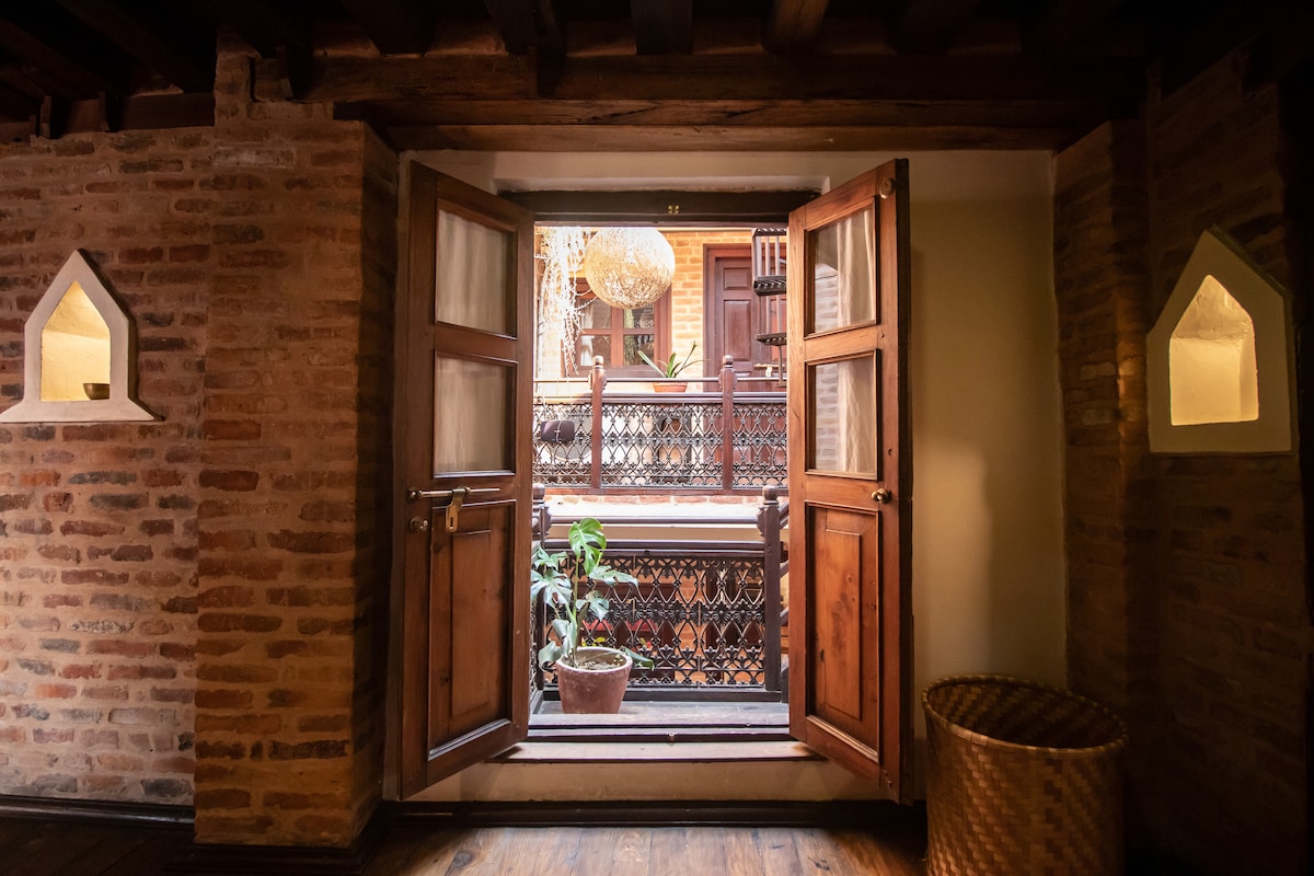 Two wooden doors are opened wide, inviting natural light into the space. A potted plant is visible on the balcony outside, while the intricate railing and interior brick walls provide a glimpse of the traditional architecture.