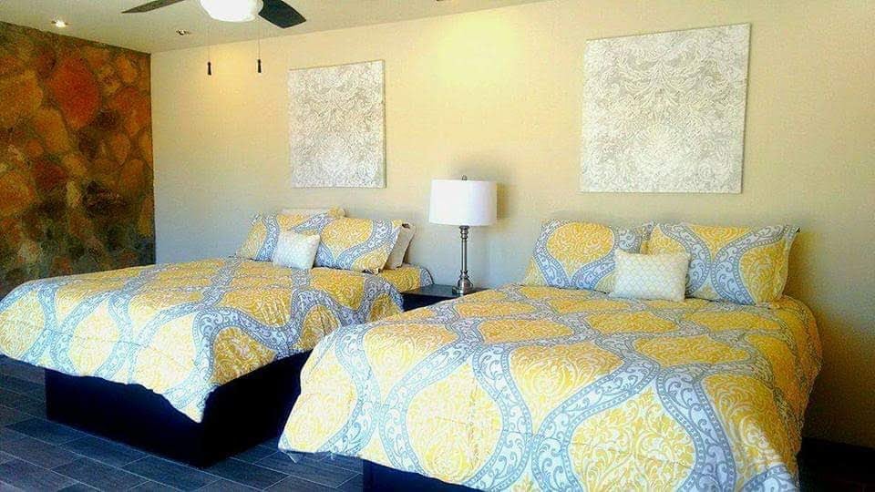 A bright sleeping area is equipped with two spacious queen beds, each dressed in yellow and gray patterned bedding. Soft decorative pillows are arranged atop the beds. A stylish lamp stands on a bedside table, providing a warm glow to the room's neutral-colored walls.