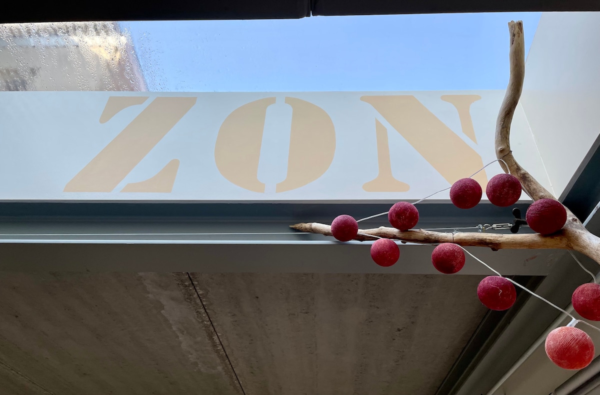 A view of the ceiling features the word 'ZON' in bold letters, with natural light streaming through a window above. Decorative red circles hang from a branch positioned near the window, adding a touch of warmth to the space.