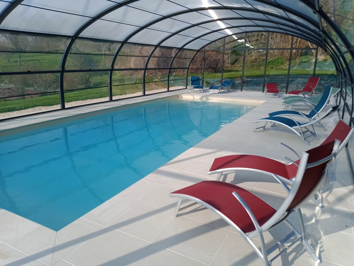 An indoor heated swimming pool is featured under a transparent canopy, with clear blue water reflecting the surrounding greenery. Several lounge chairs are positioned around the pool, showcasing a combination of blue and red colours for guest comfort.