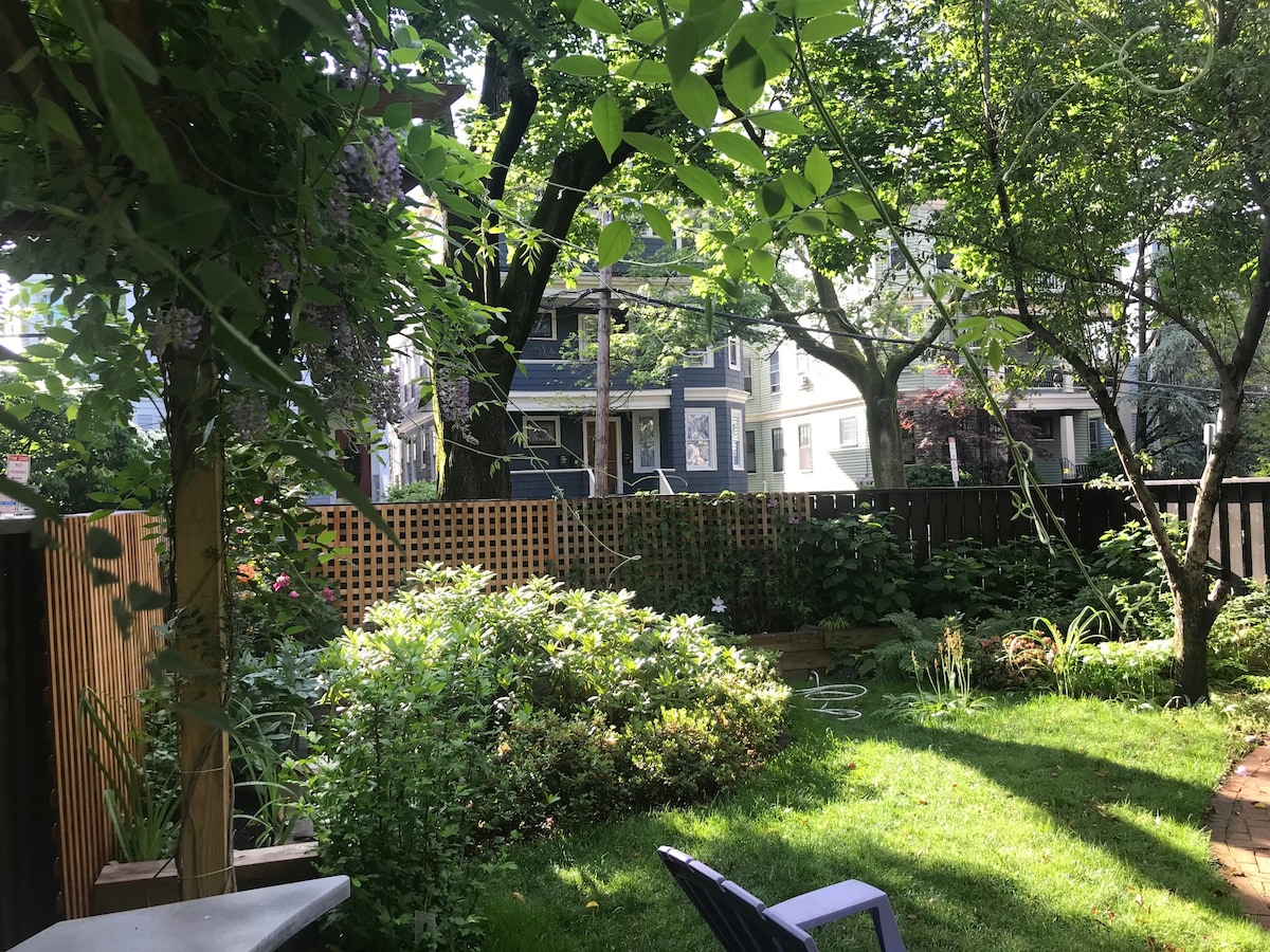 A garden space is surrounded by lush greenery and various plants, including flowering shrubs. A lawn area is visible, complemented by a wooden fence that adds privacy. Trees create a natural canopy, providing shade along with a cozy seating area featuring adirondack chairs.