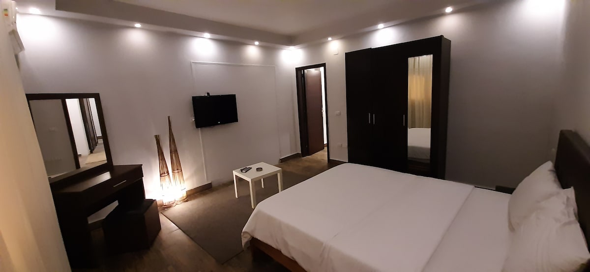 A spacious bedroom is seen, featuring a comfortable queen-sized bed with crisp white linens. A large wardrobe stands nearby, along with a flat-screen TV mounted on the wall. A minimalist desk and chair are placed beside a small coffee table, creating a functional layout.