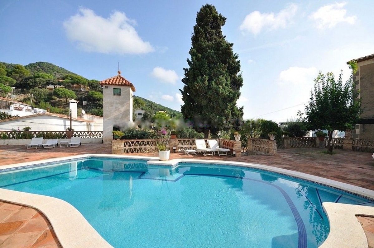 A serene pool area is surrounded by a spacious patio and lush greenery. Comfortable lounge chairs are positioned near the water, while a tall tree casts gentle shade. The backdrop features a charming Mediterranean-style building and distant hills under a clear blue sky.