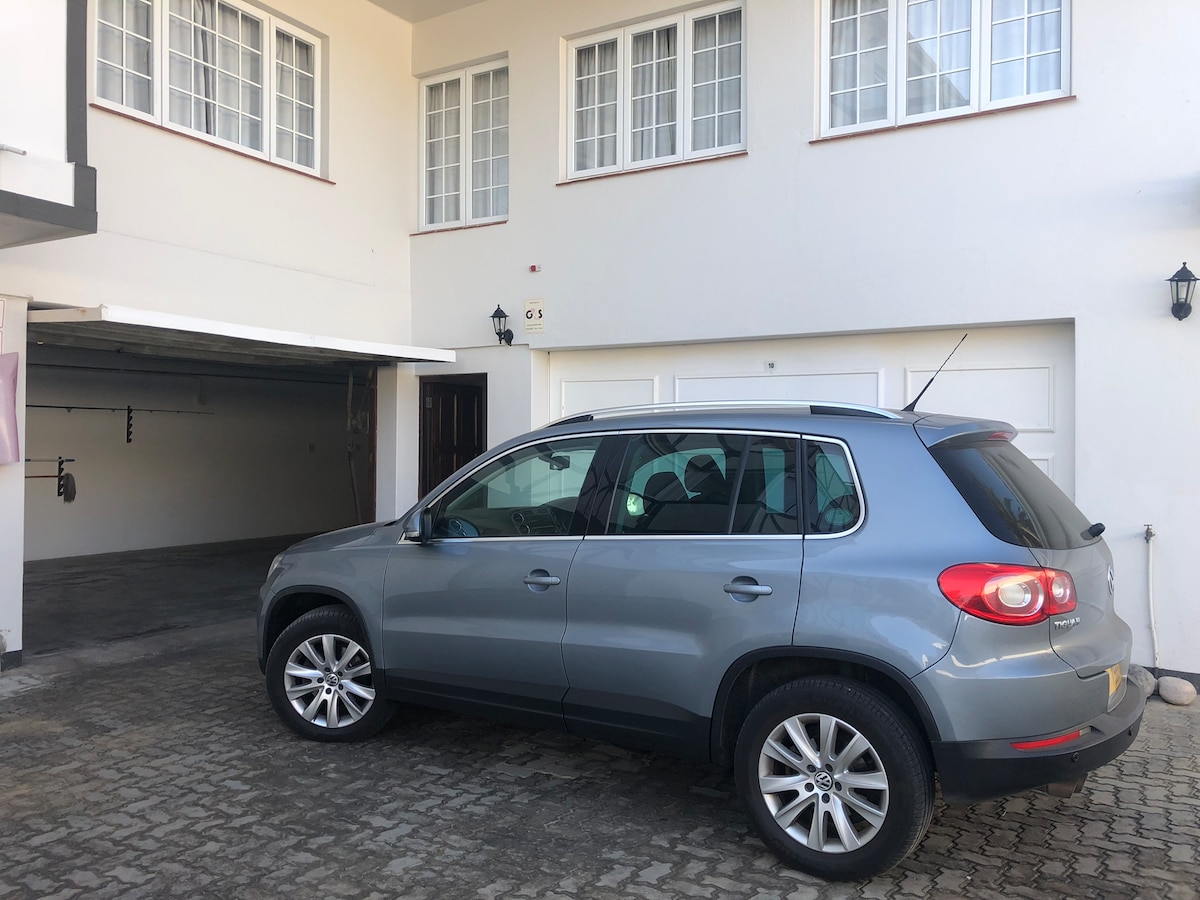 A silver SUV is parked outside a garage in front of a two-story duplex apartment. The building features large, vertical windows, allowing ample natural light. The driveway has a textured tiling, and a garage door is visible on the left side of the image.