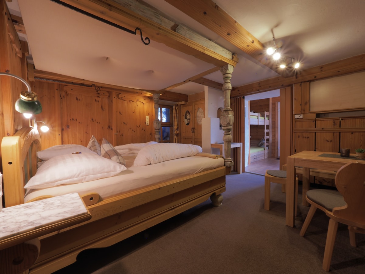 A cozy bedroom features a spacious wooden bed with neutral linens, complemented by warm wooden paneling throughout. A small dining table with chairs is positioned nearby. Soft lighting from a lamp illuminates the room, creating a calm retreat. Doorways lead to other areas of the accommodation.