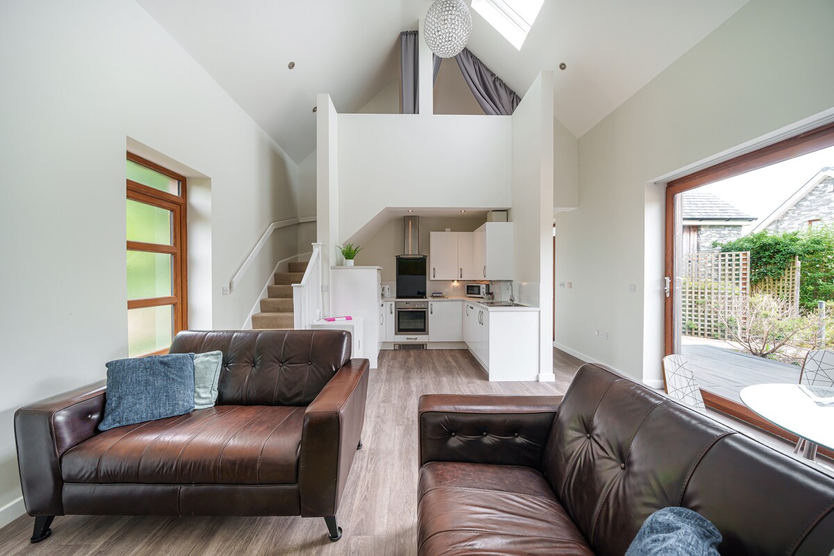 An airy open-plan living area showcases a modern kitchen with sleek white cabinetry and stainless steel appliances. Two comfortable sofas face each other, creating an inviting seating arrangement. Large windows allow natural light to fill the space, enhancing the overall brightness of the room.