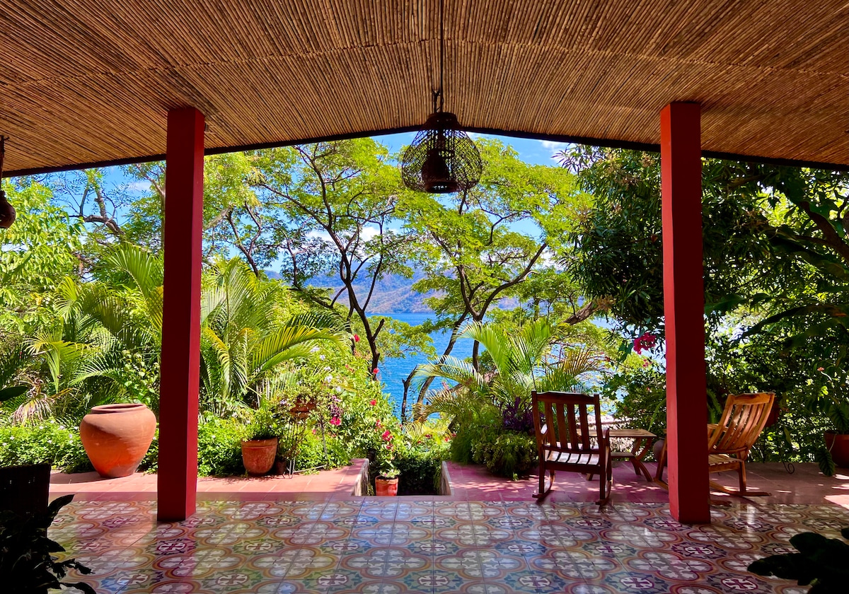 A covered terrace is framed by vibrant greenery, offering views of the tranquil water beyond. Two wooden chairs and a large earthen pot are positioned on a decorative tiled floor, which is enhanced by beams overhead. Natural light fills the space, creating a serene atmosphere.