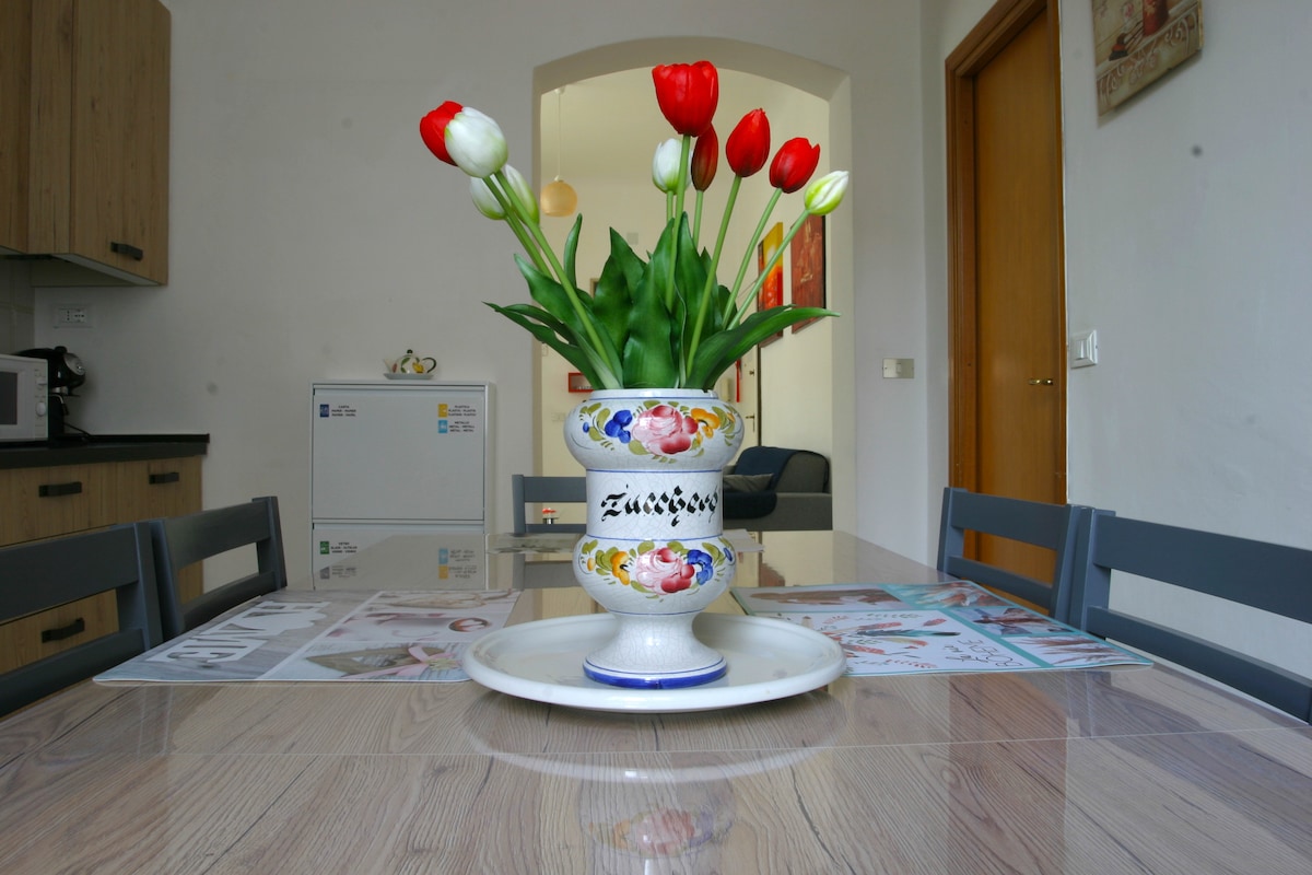 A dining table is centered in the image, featuring a decorative vase filled with vibrant red and white tulips. The table is surrounded by grey chairs, with natural light reflecting off the wooden surface. In the background, kitchen cabinetry and a refrigerator are discernible.