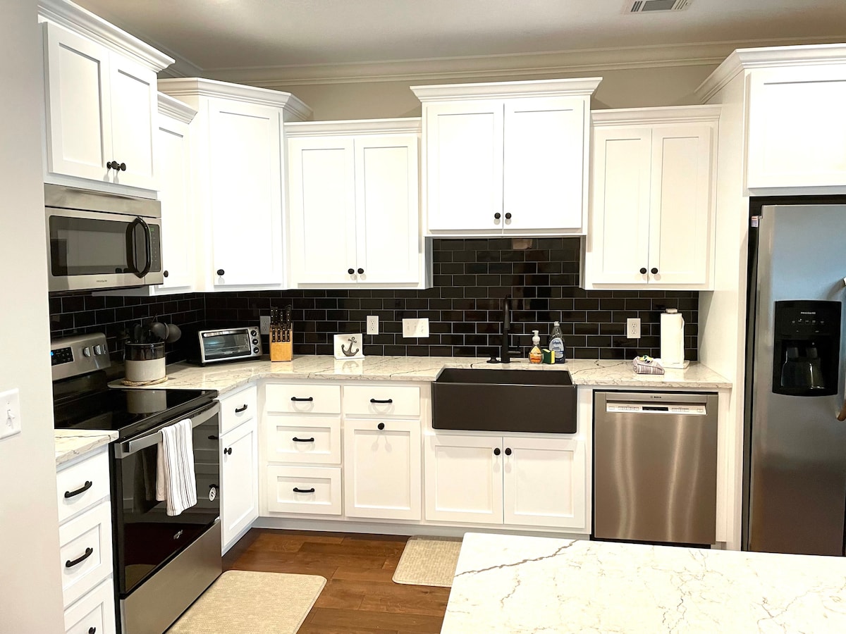 The kitchen features sleek white cabinetry paired with dark backsplash tiles. Stainless steel appliances, including an oven, microwave, and dishwasher, are included. A farmhouse sink is prominently situated in front of a window, with spacious marble countertops offering ample workspace.