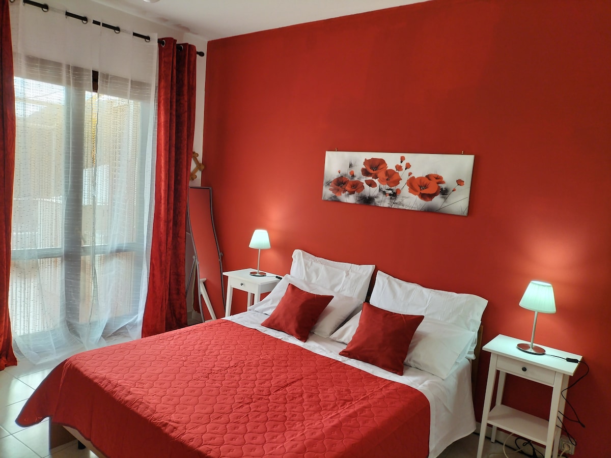 A warm-toned bedroom features a spacious bed dressed in a red quilt, accompanied by two bedside tables with lamps. Soft curtains drape over the window, allowing natural light to fill the room. A floral artwork is displayed above the bed, enhancing the inviting decor.