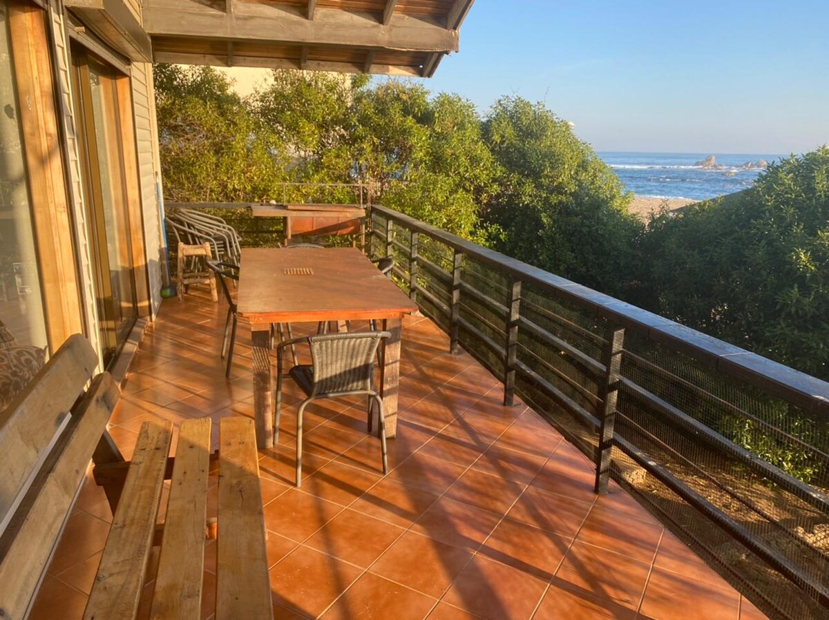 A sunlit terrace features a wooden dining table surrounded by black wicker chairs, with a view of the ocean visible beyond a black railing. Wooden benches line one side of the space, and the flooring is tiled in warm colors.