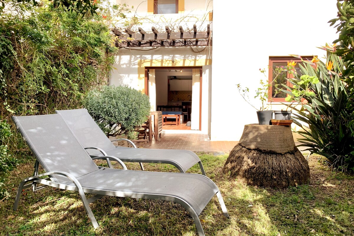 Two loungers are positioned in a serene garden with lush greenery. A natural material table stands nearby, surrounded by various plants. The entrance of the duplex is visible in the background, offering access to the indoor living space.