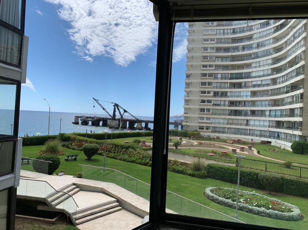 A view from an interior window reveals landscaped green areas with neatly arranged gardens and seating. In the distance, a pier is observed extending into the water, while modern buildings are visible along the shoreline under a clear blue sky.