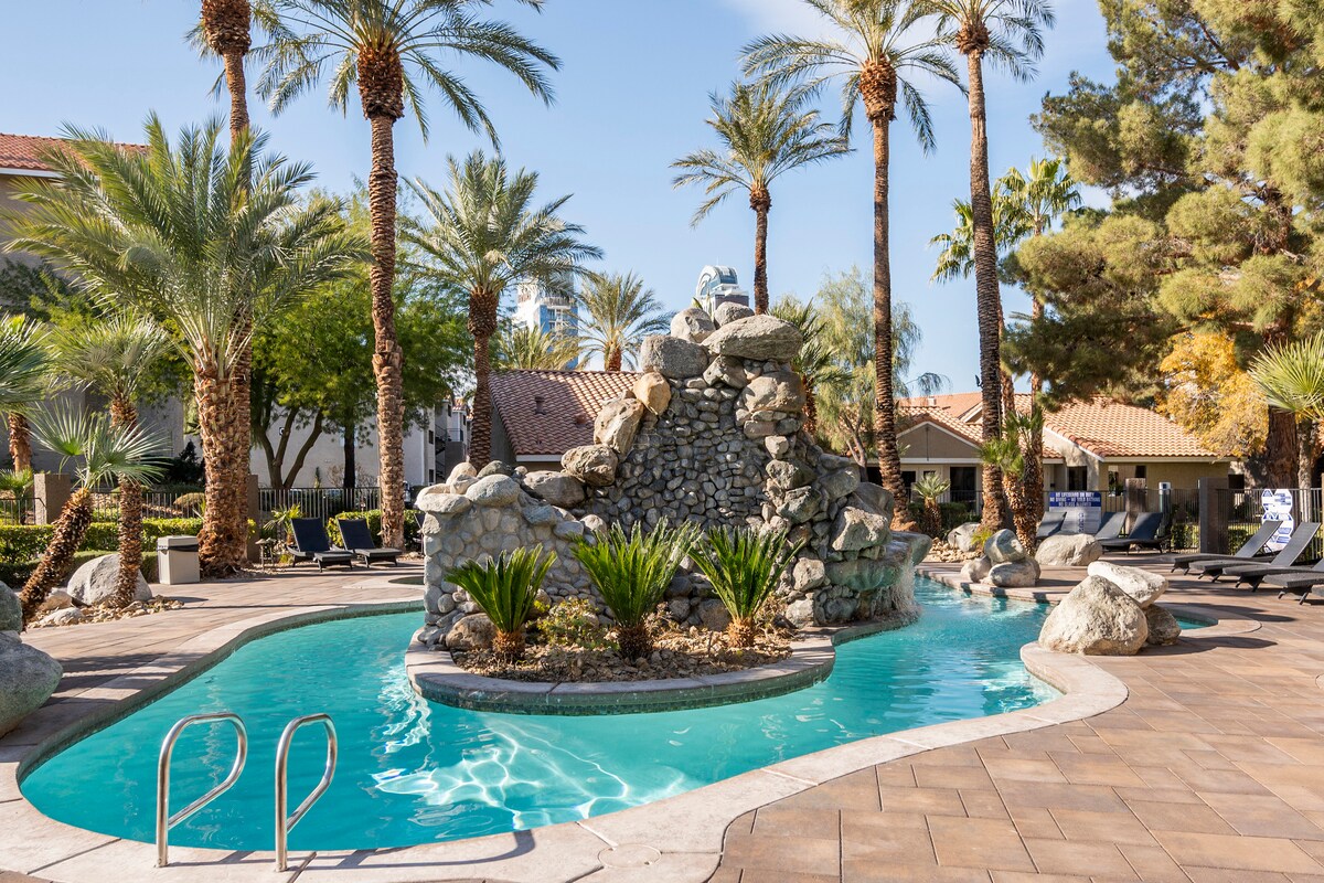 A tranquil swimming pool area is surrounded by palm trees and elegant landscaping. A rock formation creates a serene waterfall feature, while lounge chairs are placed along the pool's edge. The inviting blue water reflects the clear sky above.