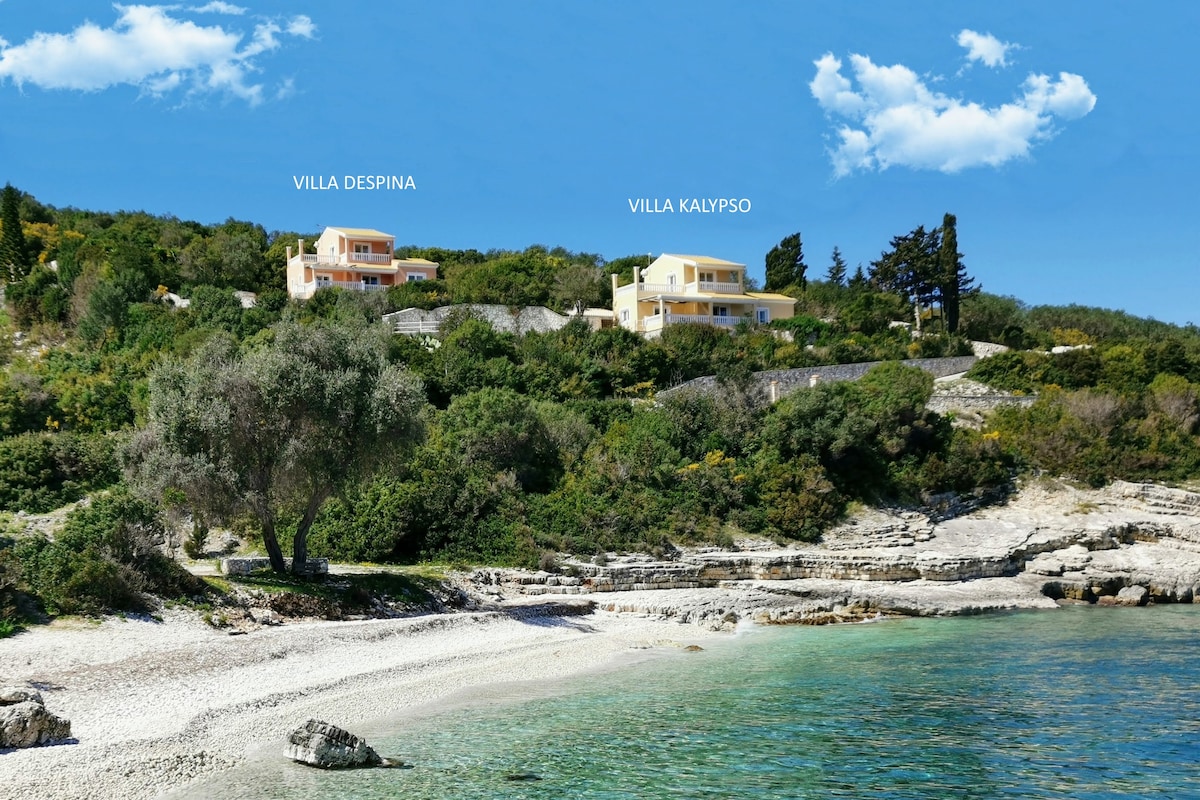 Two villas are set on a hillside overlooking clear waters. Villa Despina and Villa Kalyipso are visible, surrounded by lush greenery and rocky coastline. The sandy beach extends along the shoreline, framed by gentle waves lapping at the shore under a bright blue sky.