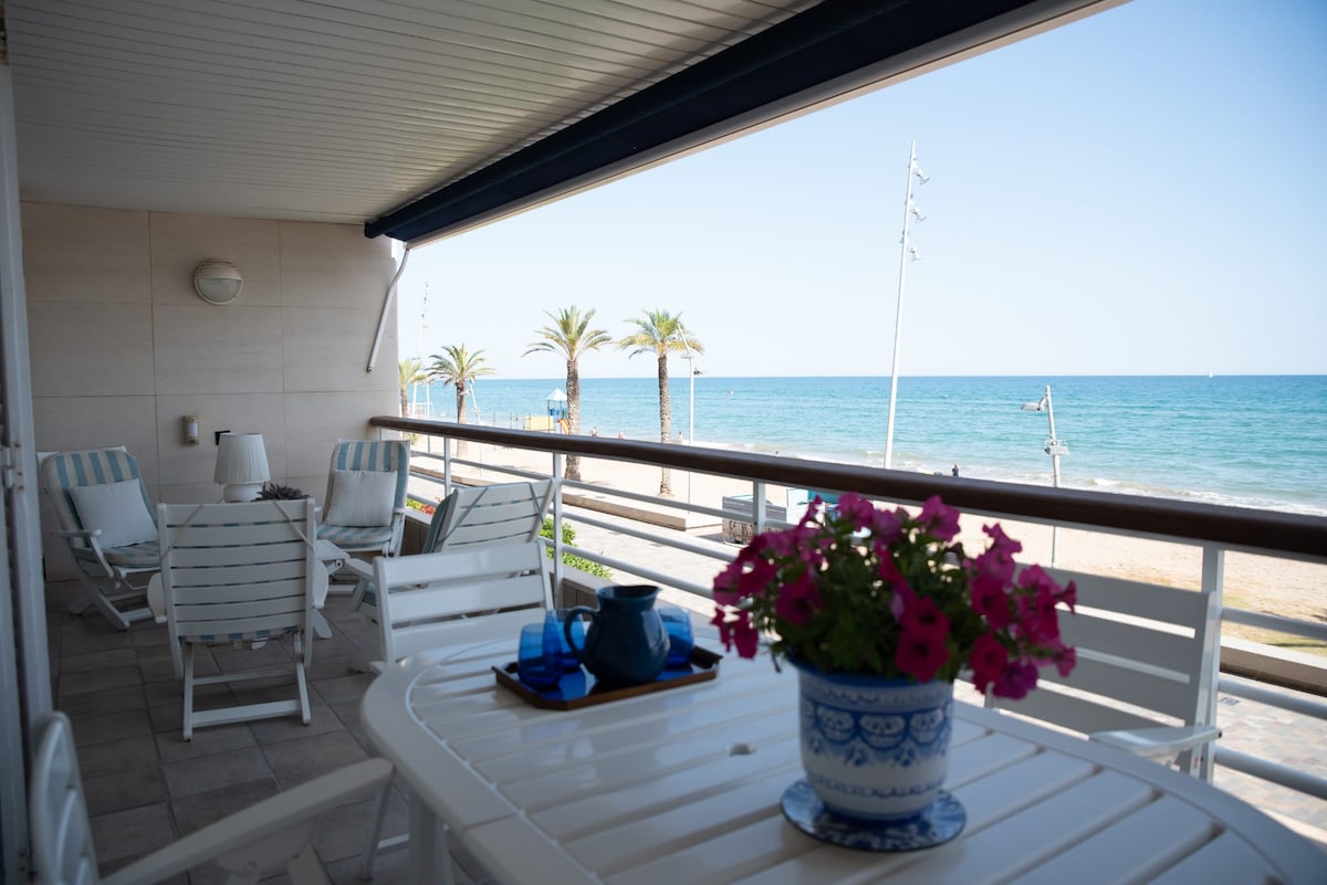 A spacious terrace is showcased, featuring a dining table surrounded by white chairs. Potted pink flowers add a touch of color. The view overlooks the beach and the sea, with palm trees lining the waterfront and a clear blue sky in the background.