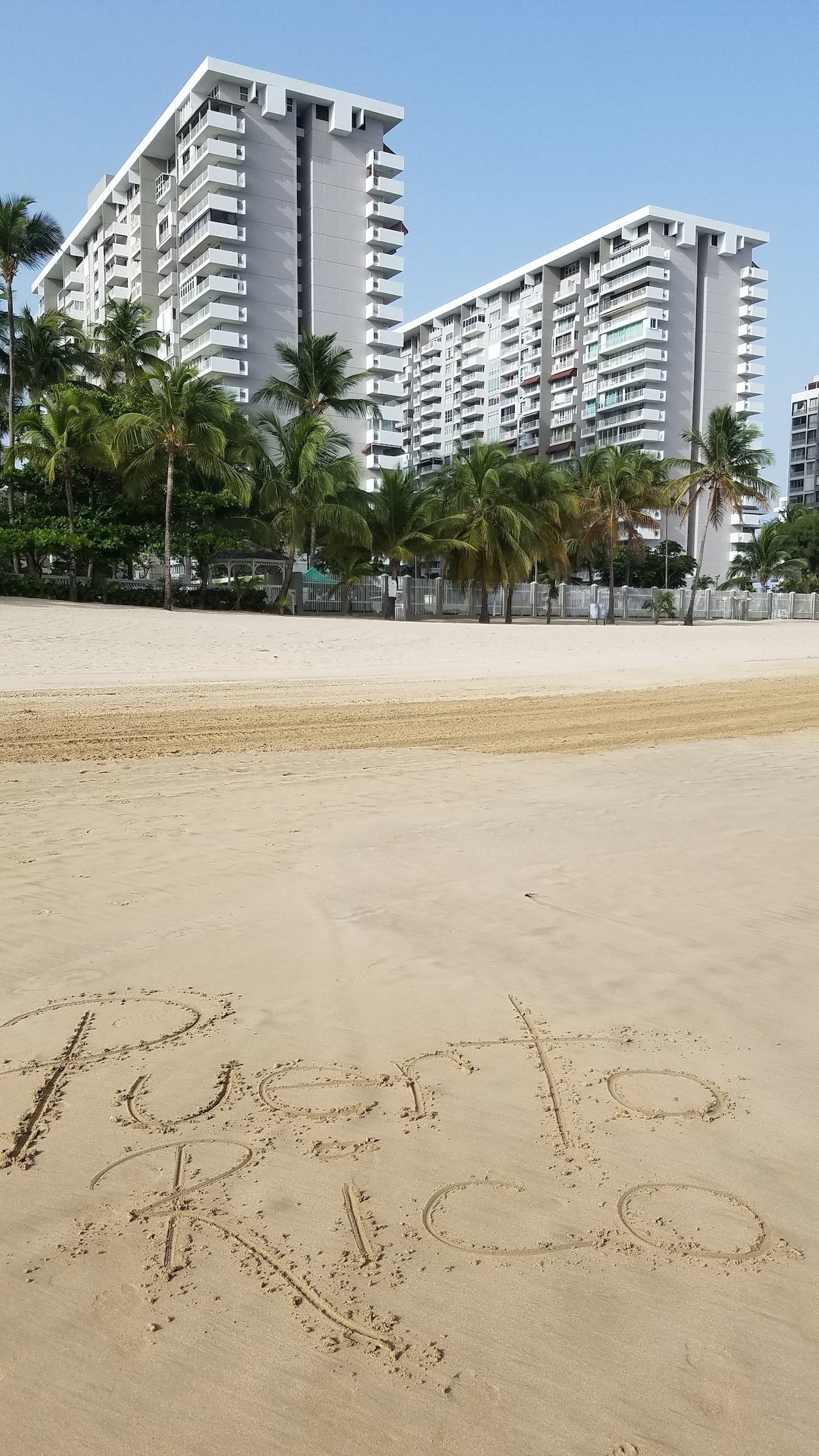 The image captures a sandy beach with the words 'Puerto Rico' drawn in the sand. In the background, tall buildings rise among palm trees under a clear blue sky, highlighting a tropical setting.