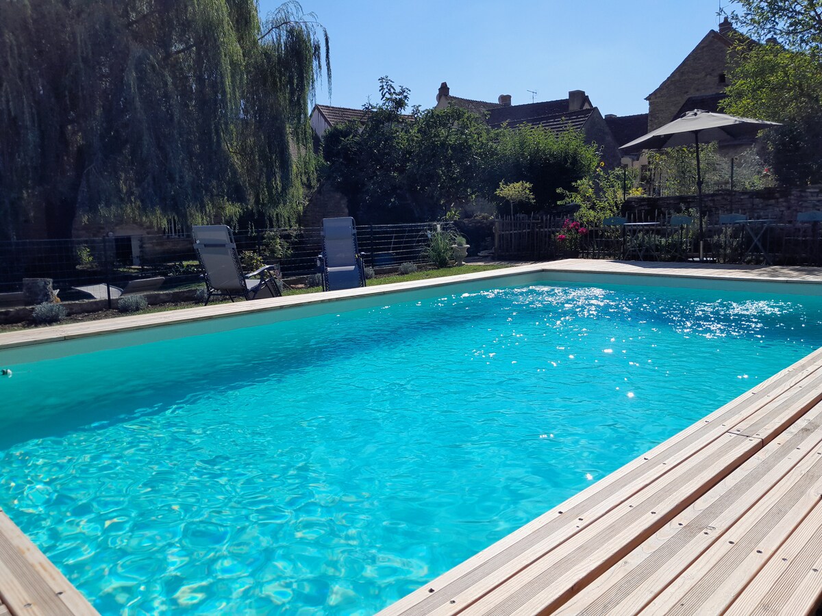 A clear swimming pool reflects the blue sky, surrounded by a wooden deck. Lounge chairs are positioned near the water, and greenery is visible in the background, creating a relaxing environment for leisure.