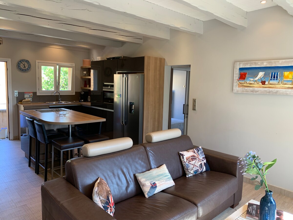 A modern living space features a comfortable brown leather sofa adorned with decorative pillows. A stylish dining area is situated nearby, with a large wooden table and high-backed chairs. The kitchen is equipped with sleek cabinetry and stainless steel appliances, accented by natural light from windows.