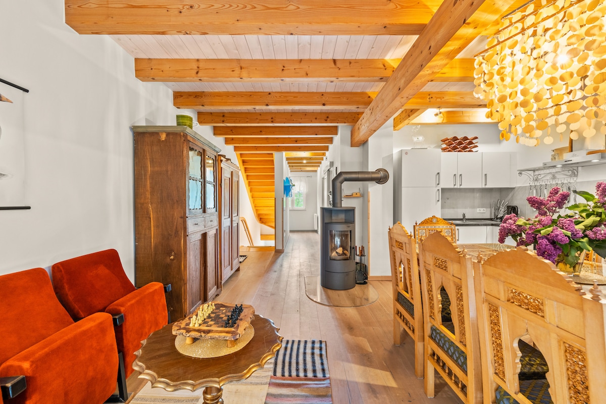 A spacious living area features exposed wooden beams and a wooden floor. A red armchair sits beside an ornate dining table, which is surrounded by intricately designed chairs. A modern wood stove adds warmth, while a colorful bouquet enhances the inviting atmosphere.