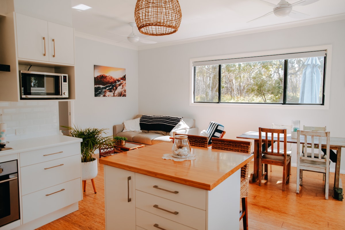 An open-plan kitchen and dining area is presented with natural light streaming through a large window. A wooden island with a light countertop is complemented by a well-equipped kitchen. A cozy seating area with a large sofa and various dining options completes the inviting space.