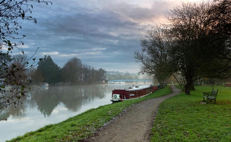 A serene riverside scene features a winding path alongside the Thames, bordered by trees and light fog. Several boats are anchored on the calm water, with a gentle reflection of the overcast sky. A wooden bench is positioned to offer a view of the landscape.