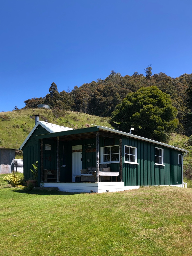 A charming cottage features a green exterior and a welcoming porch with seating. Surrounded by lush grass, the setting is framed by trees and rolling hills. Large windows allow natural light to fill the interior, showcasing the rustic character of the building.