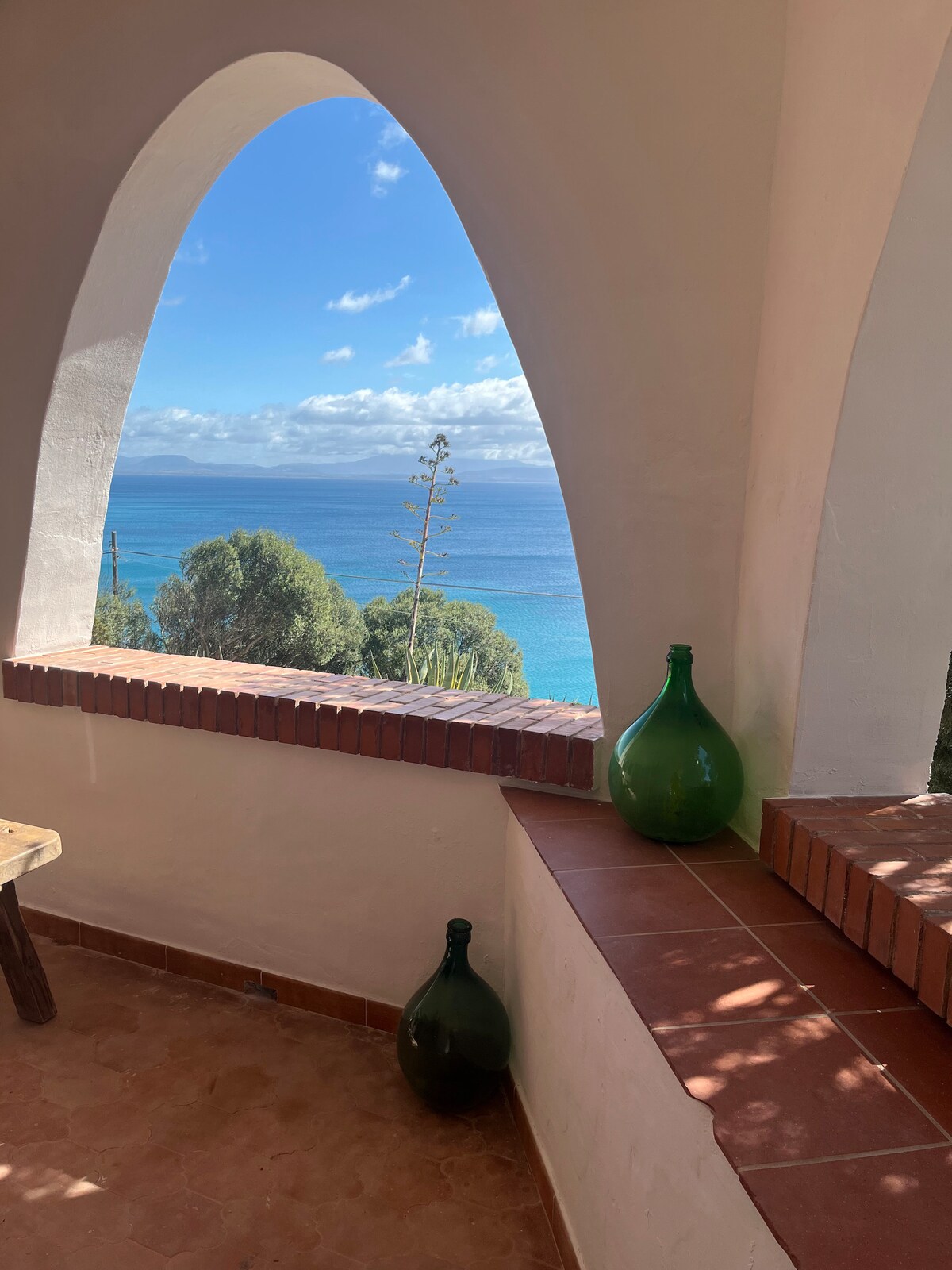 An arched window frames a stunning view of the blue sea, with greenery visible in the foreground. Two decorative green glass bottles rest on a tiled ledge, highlighting the traditional architecture and inviting scenery.