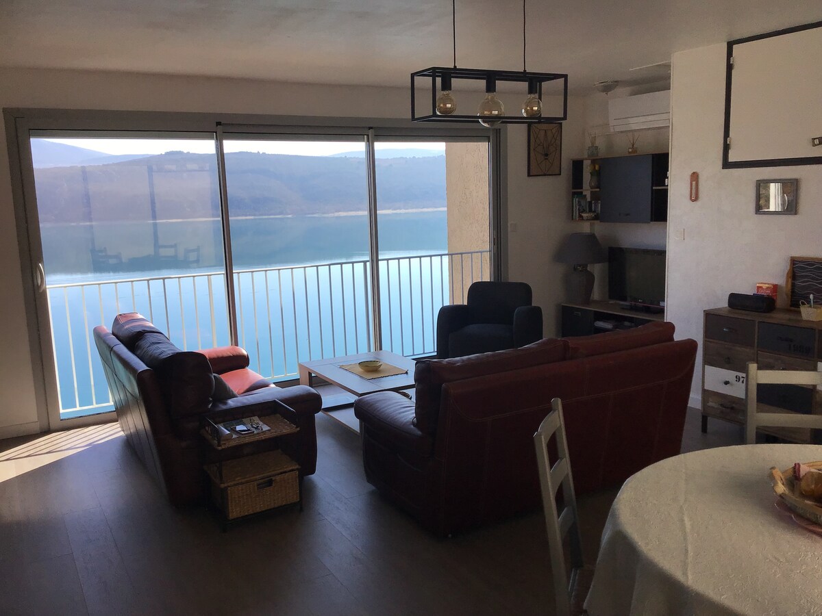The living area features large glass doors that open to a stunning lake view. A combination of leather sofas and armchairs create a comfortable seating arrangement. A round dining table sits nearby, complemented by decorative elements on the walls.