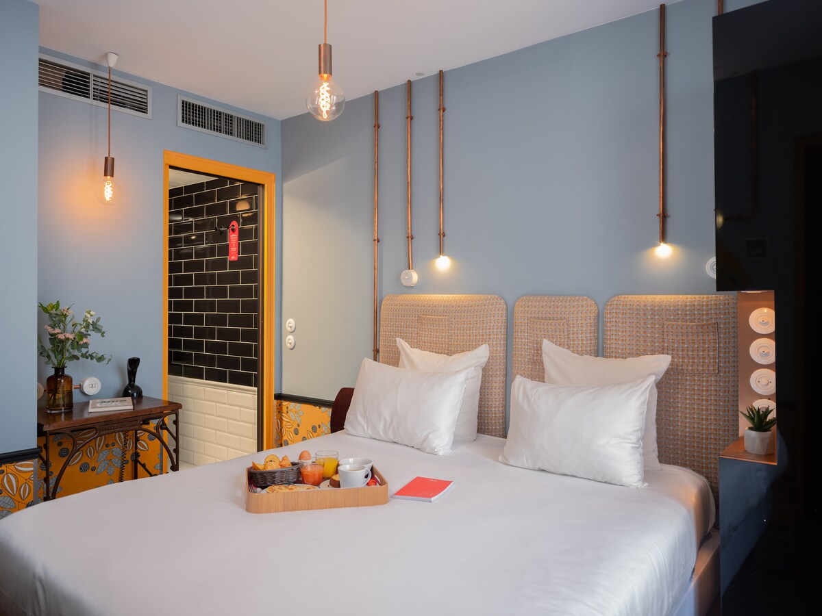 A spacious bedroom features a comfortable bed with white linens and two decorative pillows. Soft lighting is provided by hanging fixtures on either side. A tasteful kitchenette is visible in the background, complemented by a small table showcasing a fruit basket and a reading material.