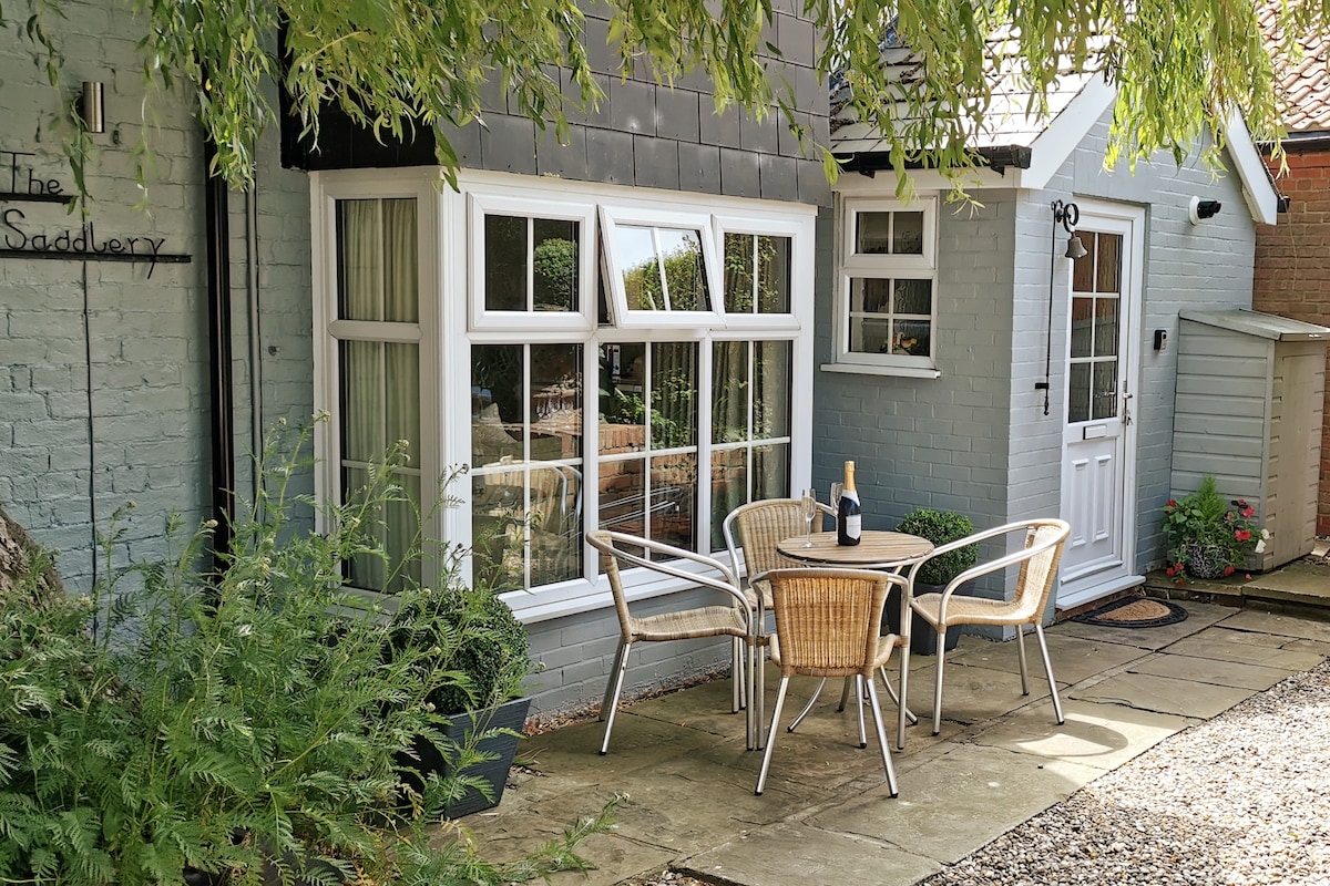 An outdoor patio area is presented with a set of four wicker chairs surrounding a round table. A bottle of champagne is placed on the table. The cottage's exterior features a combination of gray and light blue brick, with large windows allowing natural light to enter.