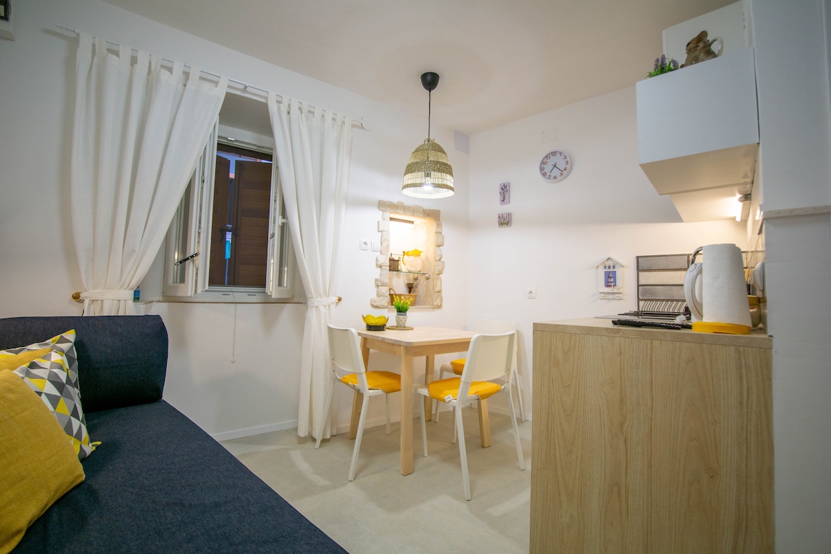 A light-filled kitchen and dining area showcases a wooden table with four yellow-accented chairs. White curtains frame the window, revealing a glimpse of the outdoors. A blue sofa with patterned throw cushions adds a cozy touch, while a wall clock and decorative items enhance the space.