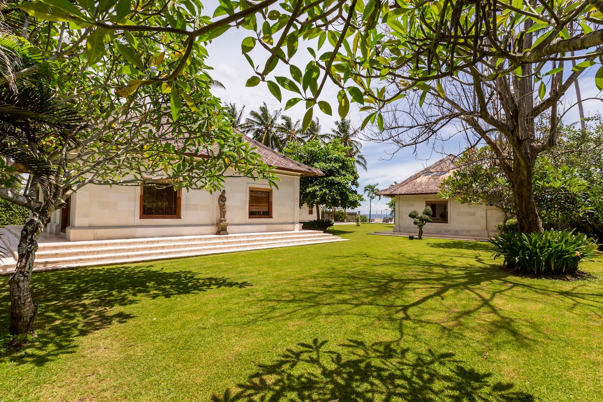 A well-maintained garden surrounds a serene villa, featuring lush green grass and ornamental trees. The architectural elements include two separate buildings, each with traditional Balinese roofs, providing a spacious and tranquil environment. Sunlight filters through the leaves, creating relaxing shadows on the ground.