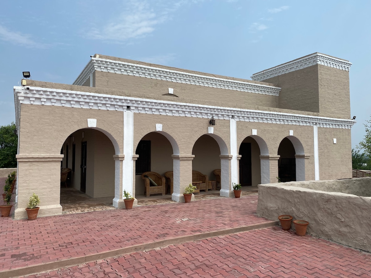 A spacious terracotta pathway leads to a charming two-story structure with distinctive arched entrances. Soft beige walls and decorative white trim highlight the architectural features. Potted plants are positioned at the base of the arches, contributing to the welcoming appearance of the exterior.