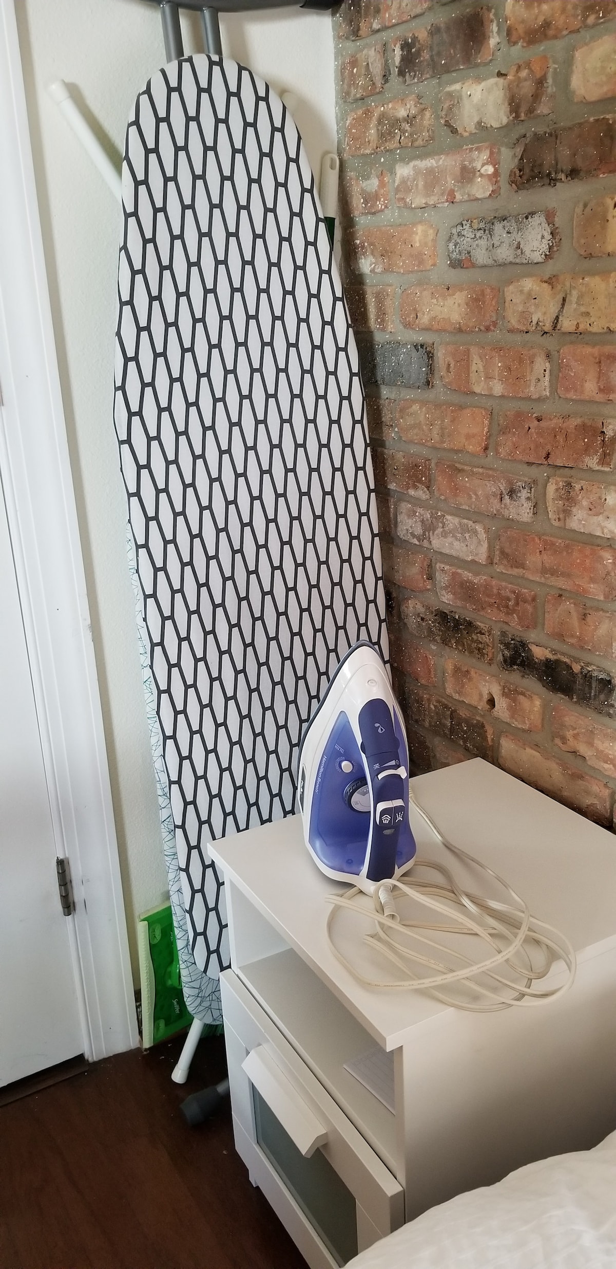 An ironing board is set up against a textured brick wall, featuring a honeycomb pattern cover. A steam iron rests beside it on a small bedside table, which includes a drawer for storage.